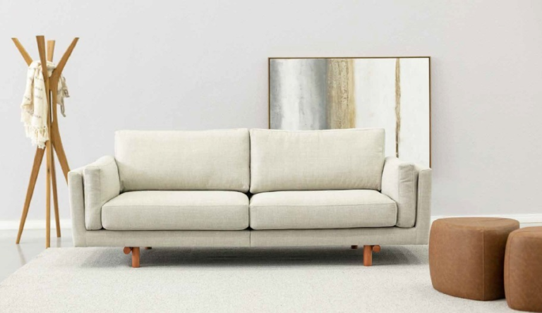 Sofá de três lugares em tom bege claro, com design contemporâneo e linhas retas, apoiado sobre pés de madeira, em sala de estar minimalista com tapete neutro, pufe em couro marrom e decoração moderna - ideal para quem busca conforto, versatilidade e elegância na escolha do sofá novo.